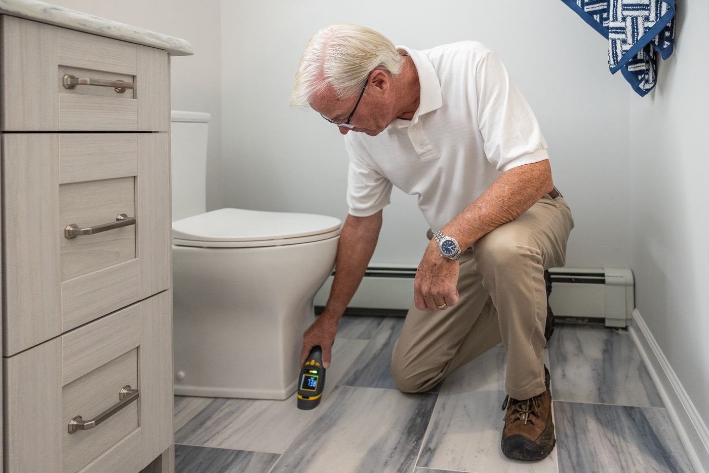Illustrative image: technician kneeling to take a moisture reading at the base of a bathroom wall